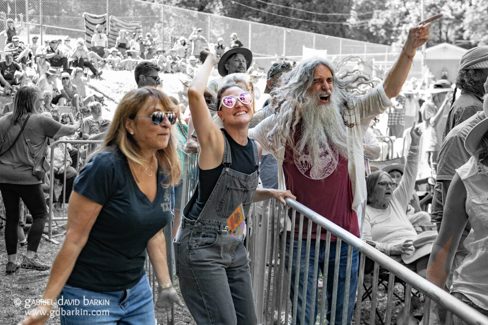 Crowd behind metal barrier at an outdoor event; a smiling woman with heart-shaped pink sunglasses raises her arms beside a bearded man cheering with his fist raised.