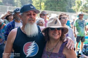 Smiling couple at an outdoor festival; man with a gray beard wearing a dark tank top and cap, woman in a wide-brim hat with a pine tree design, surrounded by a cheering crowd.