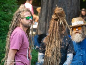 Three people with long dreadlocks standing together at an outdoor event; one man in a pink shirt wearing green mirrored sunglasses, another person in a hat and lace sunglasses, and a third with tattooed arms partially visible.