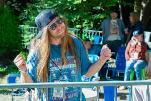 Man with long hair, backwards cap, and sunglasses posing playfully at an outdoor event. A metal barrier and other people are in the background.
