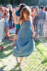 Woman in a denim dress dancing barefoot at an outdoor crowd behind a metal barrier.