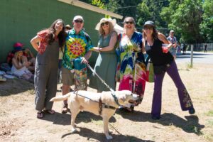 Group of people posing together outdoors in bright tie-dye clothing, with a tan dog on a leash in the foreground.