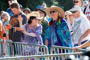 Group of people behind barricades at an outdoor event; a woman in a purple tie-dye outfit dances with hands raised.
