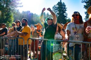 Crowd of people behind metal barriers at an outdoor event, some with sunglasses and hats, raising their arms as sunlight filters through trees.