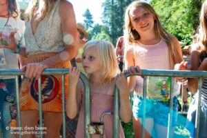 Children lean on a metal barrier at an outdoor festival, sunny day with green trees in the background.