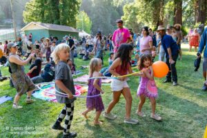 Crowd of festival attendees dancing and socializing on a sunny grassy field in a park, with children playing with hula hoops in front.