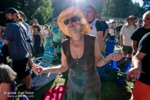 Crowd at an outdoor event; central woman wears a large straw hat and sunglasses among festivalgoers on a sunny day.