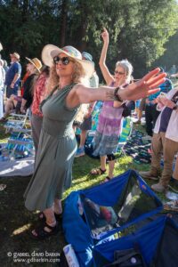 Sunlit outdoor crowd waving arms and cheering at a park event, with folding chairs and trees in the background created by bright summer sun.