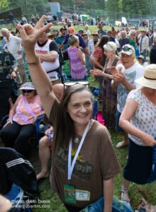 Crowd of people at an outdoor festival; foreground woman in a brown shirt smiles and raises her arm toward the camera.