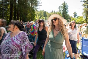 Crowd of smiling people at an outdoor festival among trees on a sunny day, wearing casual summer clothing and hats.