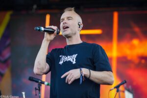 Male singer in a black T-shirt sings into a handheld microphone on a colorful concert stage.