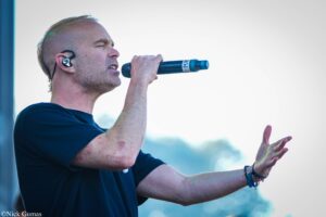 Man singing into a handheld microphone outdoors, wearing a dark T-shirt with in-ear monitor in his ear.