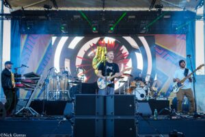 Rock band performing on stage with guitarist center and bassist right, colorful backdrop and large LED screen behind them.