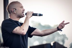 Male singer on stage, holding a microphone and singing with an in-ear monitor visible in his ear, his other hand gesturing outward during an outdoor performance.