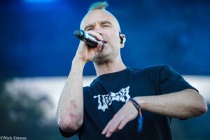 Male singer performing on stage, microphone held close to mouth, blue blurred background behind him.