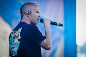 Male singer in a black T-shirt, singing into a handheld microphone on stage.