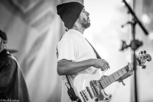 Black-and-white image of a man wearing a beanie and a white shirt playing a bass guitar on stage, captured in profile.