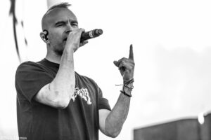 Male singer performing into a handheld microphone on stage, raising one index finger in the air.