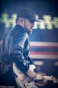 Side profile of a guitarist wearing a dark cap and jacket, playing a decorated white electric guitar with stickers on a blurred stage background.