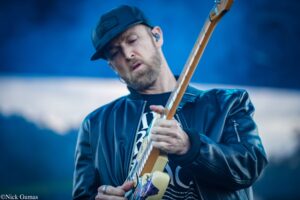 Guitarist playing an electric guitar outdoors, wearing a dark cap and leather jacket, focused on the fretboard.