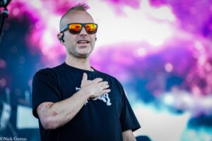 Male performer wearing sunglasses on stage, hand over chest, colorful blurred background of purple lights. (informative)