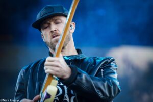 Close-up of a man playing an electric guitar on stage, wearing a dark cap and leather jacket with blue lighting in the background.