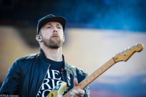 Male guitarist wearing a black cap and leather jacket, playing an electric guitar on stage. (informative)