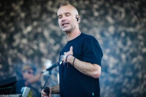 Man performing on stage, wearing a dark T-shirt and a wireless ear monitor, holding a microphone close to his mouth.