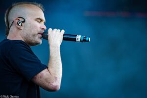 Male singer in a black shirt passionately singing into a handheld microphone with an in-ear monitor, on a dark blue stage backdrop.