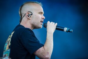 Male singer in a black T-shirt delivers a performance into a handheld microphone against a deep blue background, shown in profile with an in-ear monitor visible in the ear.