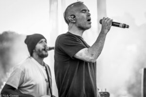 Male singer in foreground singing into a microphone on stage; eyes closed, wearing a dark T-shirt, with another musician in the background.