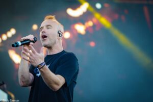 Male singer in a black T-shirt sings into a handheld microphone on stage with stage lights in the background (live performance).