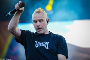 Male singer on stage, raising a microphone while eyes are closed, wearing a black T-shirt and a wireless in-ear monitor against a blue-lit background.