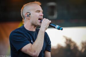 Man singing into a handheld microphone during a live outdoor performance, wearing in-ear monitors and a dark T-shirt.