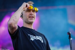 Male performer on stage adjusting yellow mirrored sunglasses during a performance, with a blue-purple backdrop and a headset in his ear