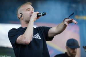 Male singer on stage, microphone held close to mouth, in-ear monitor visible, arm raised as he performs.