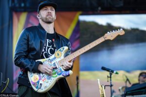 Male guitarist in a black leather jacket playing a sticker-covered white electric guitar on stage
