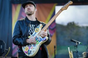 Guitarist wearing a black leather jacket and cap plays a cream-colored sticker-covered electric guitar on an outdoor stage.
