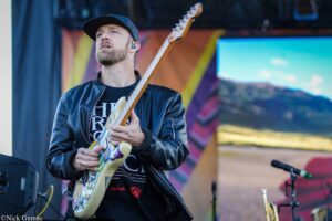 Male guitarist wearing a black cap and leather jacket, playing a sticker-covered electric guitar on stage with colorful backdrop behind him, looking upward.