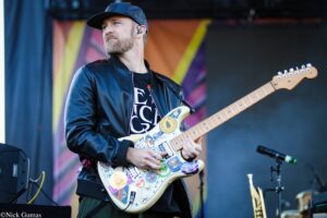 Male guitarist performing on stage, wearing a dark cap and leather jacket, playing a heavily stickered white electric guitar against a colorful backdrop.