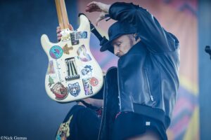 Guitarist wearing a cap and black jacket plays a cream-colored sticker-covered electric guitar on stage.