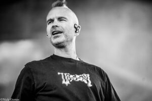 Black-and-white portrait of a man singing, wearing an in-ear monitor and a dark T-shirt, visible from the chest up outdoors.