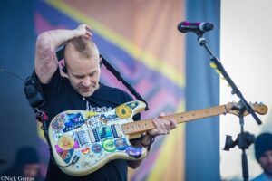 Musician on stage playing a sticker-covered electric guitar, hand resting on his head, in front of a colorful backdrop and microphone nearby.