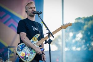 Male guitarist wearing a black shirt sings into a microphone while playing a sticker-covered electric guitar on stage.