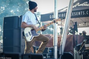 Musician wearing a beanie and white shirt playing a cream Fender bass on an outdoor stage at a festival.