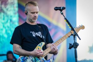 Male guitarist in a black shirt playing a decorated electric guitar on a colorful stage backdrop, wearing in-ear monitors.