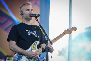Male guitarist singing into microphone on stage, wearing a black T-shirt and wireless in-ear monitor clip, guitar decorated with stickers