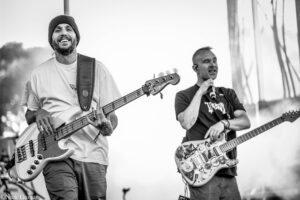 Two male musicians performing on stage: a smiling bass guitarist on the left and a vocalist with a decorated guitar on the right (black-and-white).