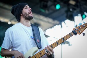 Male bassist wearing a black knit beanie and white T-shirt, playing a cream-colored Fender bass on stage under lights.