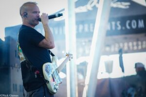 Male guitarist wearing ear monitors sings into a handheld microphone while playing a sticker-covered electric guitar outdoors under bright light.